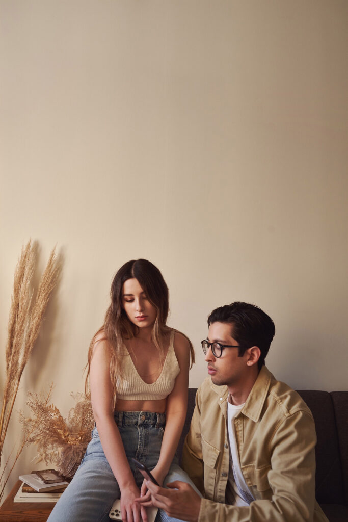 Gaby and Me are seated on a couch in a room with a beige wall. Gaby wears a white tank top and blue jeans while I wear glasses and a beige shirt. They appear to be in a relaxed setting with Me holding a remote control.
